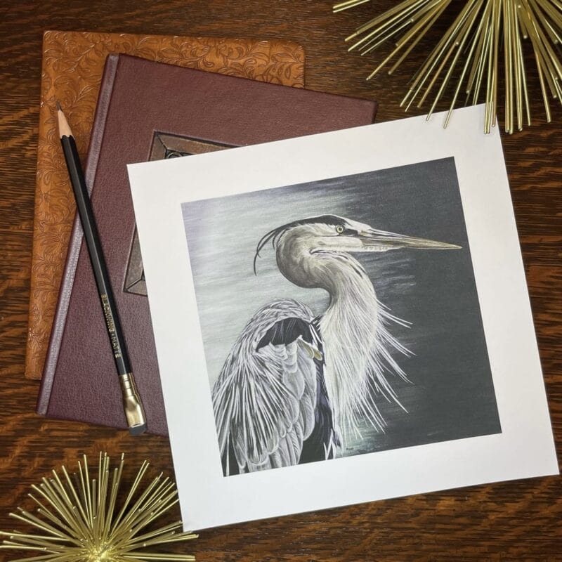 A detailed watercolor painting of a gray heron with a long beak and striking feathers is artfully placed atop two ornate books at the Holiday Bazaart 2024, accompanied by a pencil and golden decorative spikes on a wooden surface.