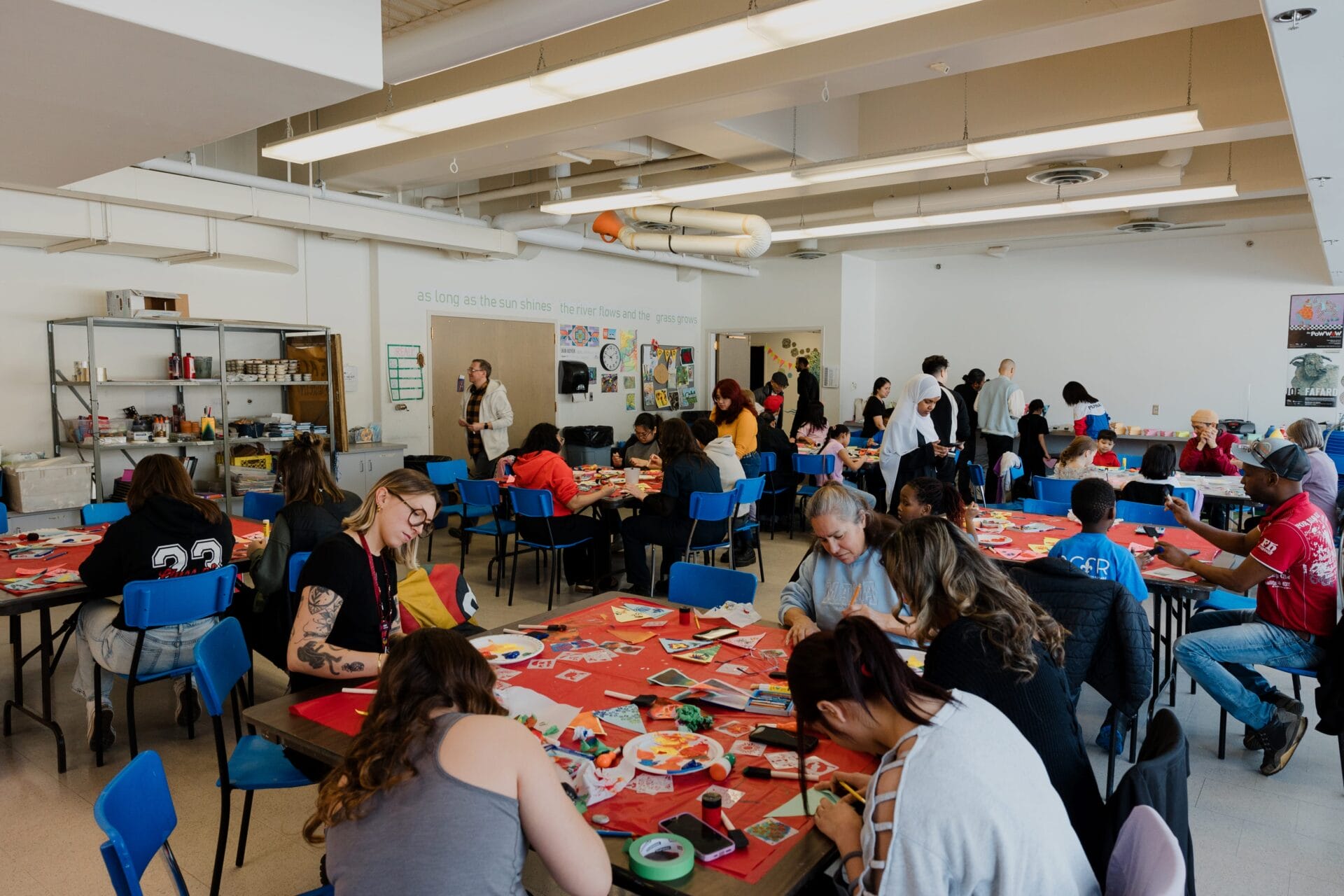 A Weekly We Make session fills the classroom, with students and adults sitting at tables engaged in various art projects. Supplies and artwork are scattered on red tablecloths as teachers assist participants in the bright, open room.