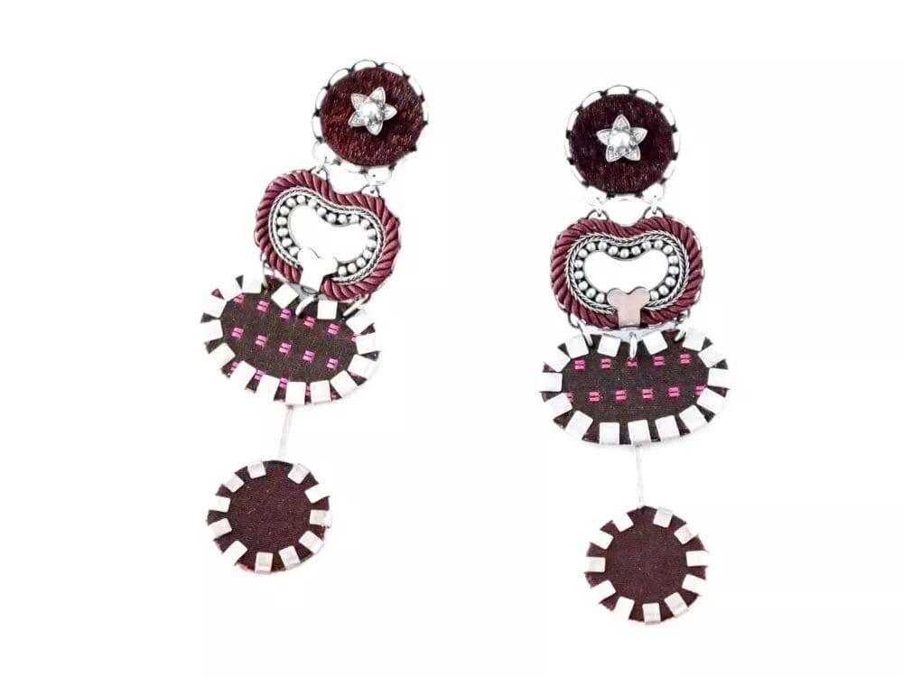 Earrings by Ayala Bar are a stunning pair of unique, handcrafted earrings featuring a mix of dark red and silver elements. These earrings boast three distinct sections: a circular top adorned with a star emblem, a middle ring showcasing intricate braided detail, and a bottom disc with an elegant patterned surface. An added touch includes detachable decorative matching studs.