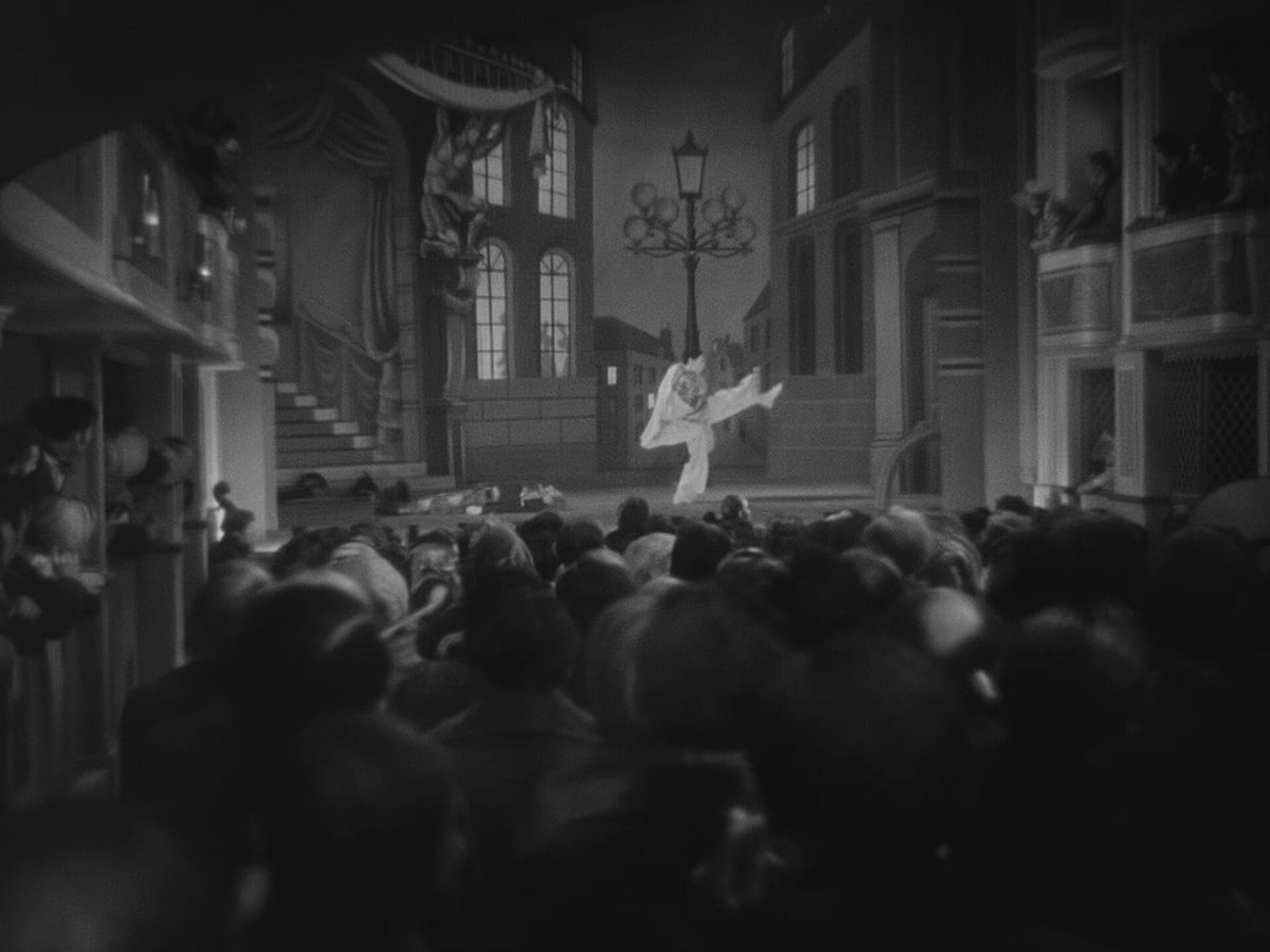 Une photographie en noir et blanc montre une scène de théâtre en salle avec un public bondé. Sur scène, un artiste semble danser ou se livrer à un acte dramatique. Le décor présente un paysage urbain détaillé, notamment des bâtiments, des fenêtres et un lampadaire.
