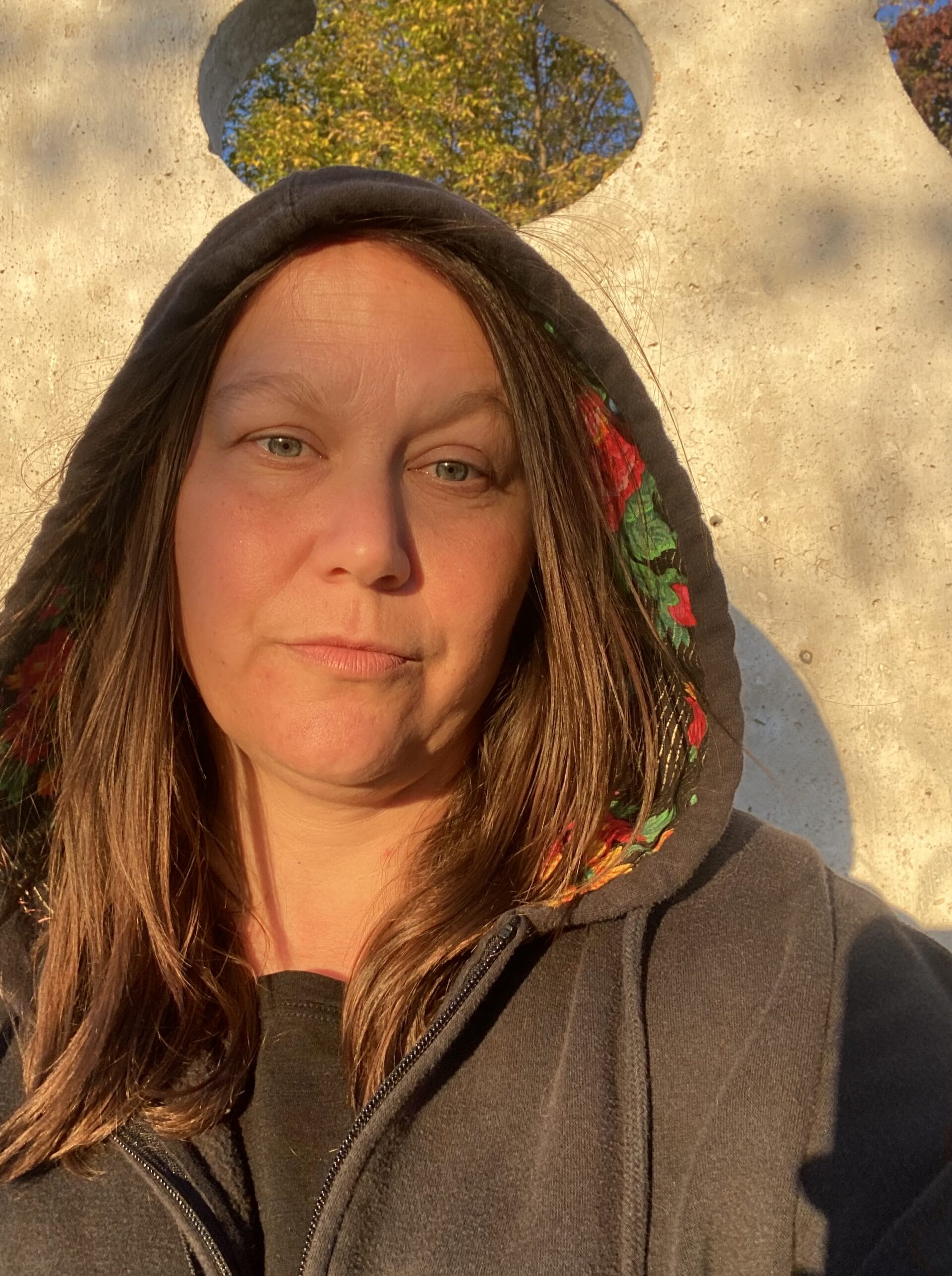 Une femme aux longs cheveux bruns porte un sweat à capuche sombre avec une doublure à motifs floraux. Elle se tient à l'extérieur, en plein soleil, devant un mur de couleur claire avec des découpes circulaires et des arbres avec des feuilles d'automne sont visibles derrière elle.
