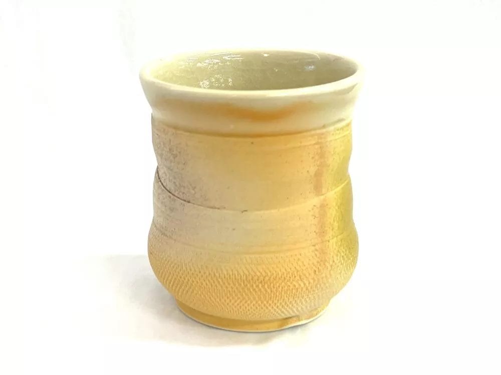 The Big Muddy-Riverhurst Meteor Cup by Jay Kimball is a small ceramic cup with a smooth, slightly flared rim and a textured curved body, glazed in warm yellow and beige tones for a beautiful gradient effect.
