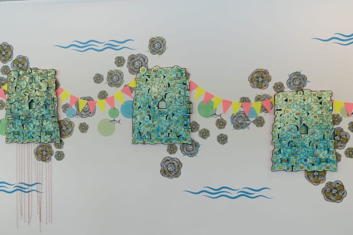 Installation artistique abstraite composée de trois panneaux texturés bleu-vert, composés de petits carrés et de découpes en forme de portes, ornés de fanions colorés. Le fond est orné de motifs floraux et de lignes bleues ondulées évoquant l'eau.