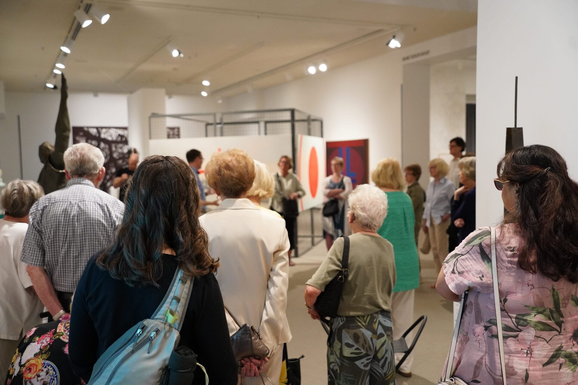 A group of people stand closely together in an art gallery, attending one of the classes & workshops, as they listen to a speaker. Various abstract artworks are displayed on the white walls in the background. Most viewers have their backs to the camera.