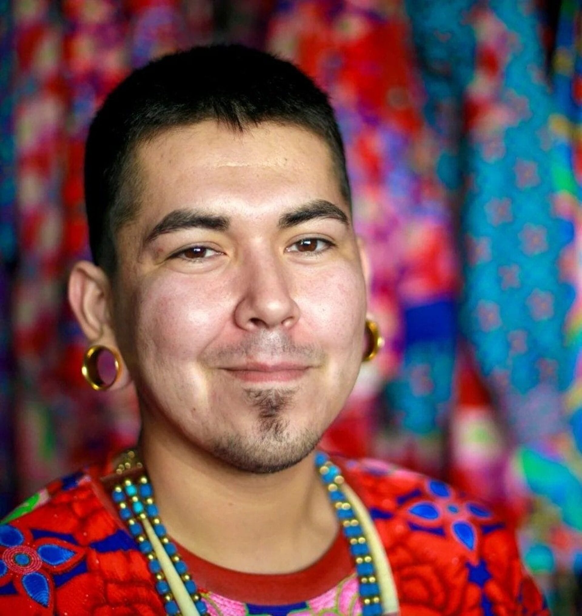 A person with short hair, wearing large gold hoop earrings and vibrant, colorful clothing with various patterns, smiles at the camera. The background is filled with similarly colorful and patterned fabric.