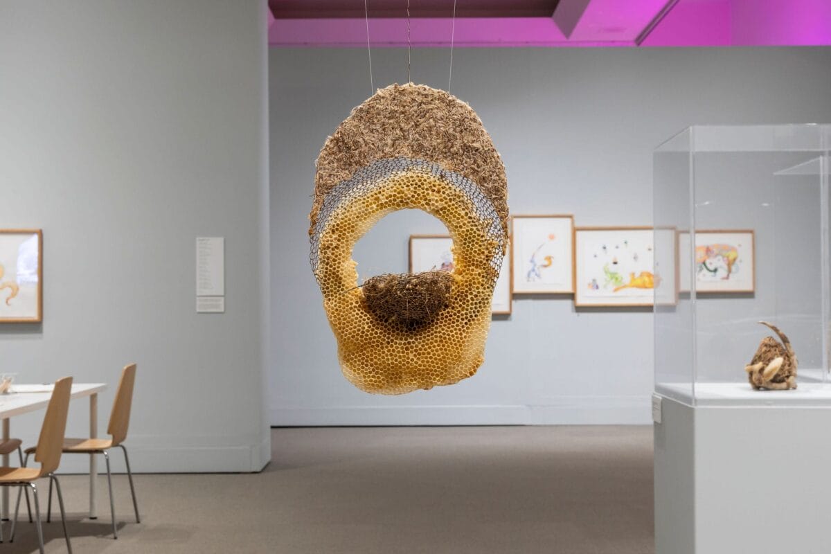 Une grande sculpture circulaire ressemblant à un nid d'abeilles est suspendue au plafond d'une galerie d'art. Des œuvres d'art encadrées et des chaises sont visibles à l'arrière-plan, avec un éclairage doux et des murs de couleur neutre.
