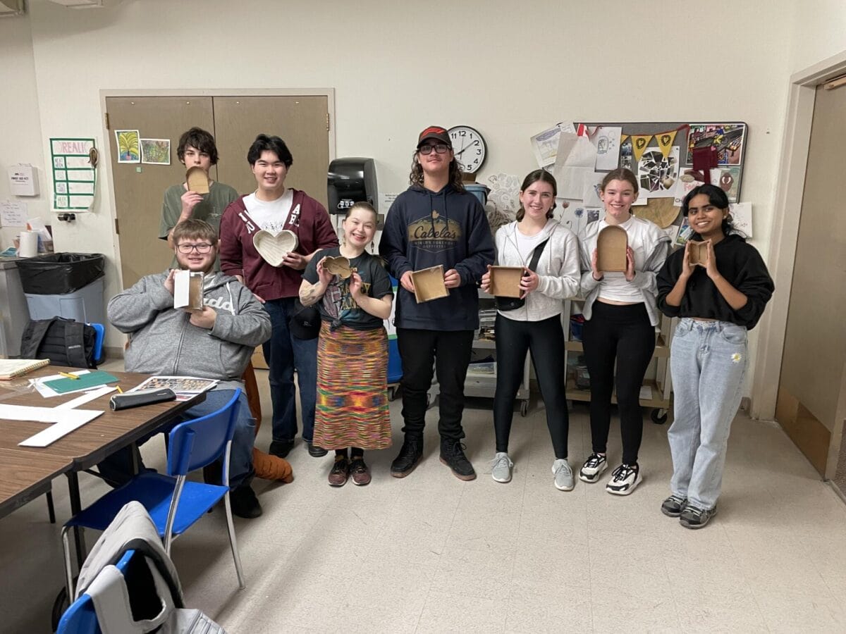 Un groupe de huit personnes souriantes se tient à l'intérieur, chacune tenant un projet d'artisanat en bois. Les projets varient en forme et en taille, y compris des cœurs et des pièces rectangulaires. Ils sont dans une pièce avec des tables, des chaises bleues et divers papiers et œuvres d'art sur les murs derrière eux.