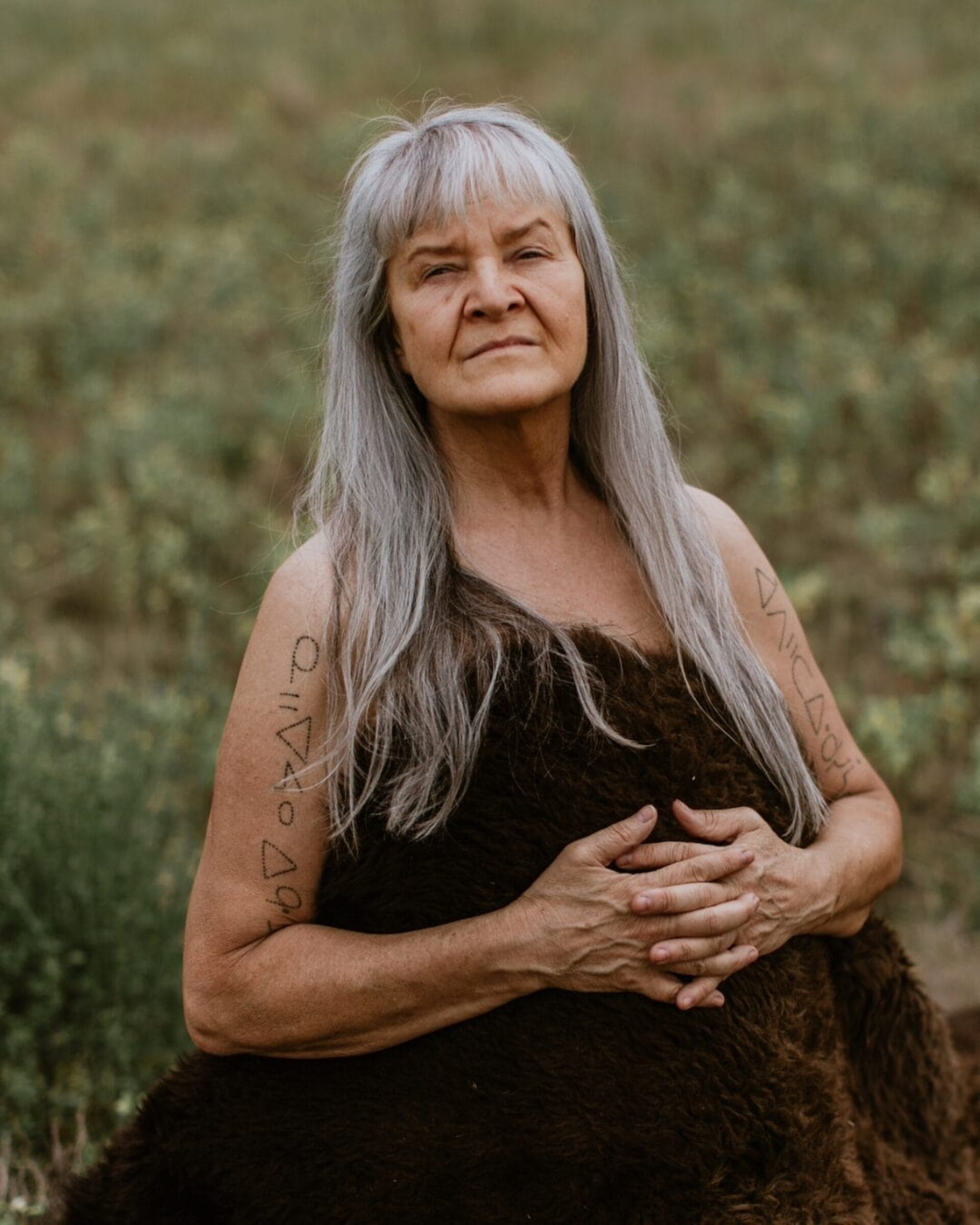 An older woman with long gray hair wears a brown fur-like wrap outdoors, standing in a green field during the Opening Reception for Indian Theatre. She has symbols drawn on her upper arms and looks confidently at the camera, hands resting on her stomach.