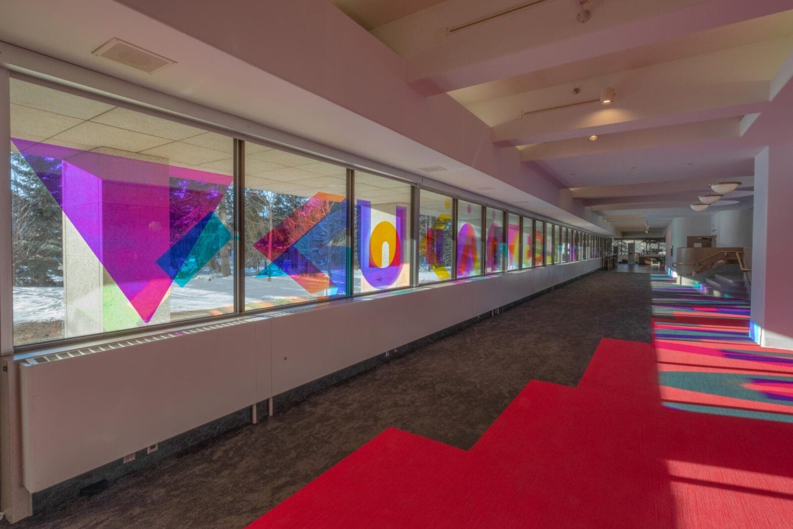 A brightly lit corridor with a long row of windows on the left side, featuring vibrant, geometric stained glass art in various shapes and colors. The sunlight casts colorful patterns onto the floor, which is covered in a mix of red and gray carpeting.