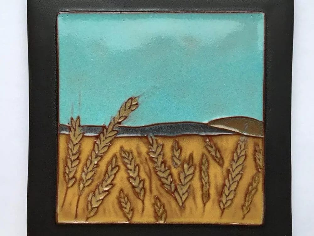 A square ceramic **Grain/Barley Tile by Brian McArthur and Dawn Detarando** features a scenic landscape with golden wheat fields in the foreground and rolling hills under a clear blue sky in the background. The tile has a dark border that frames the vibrant and textured illustration.