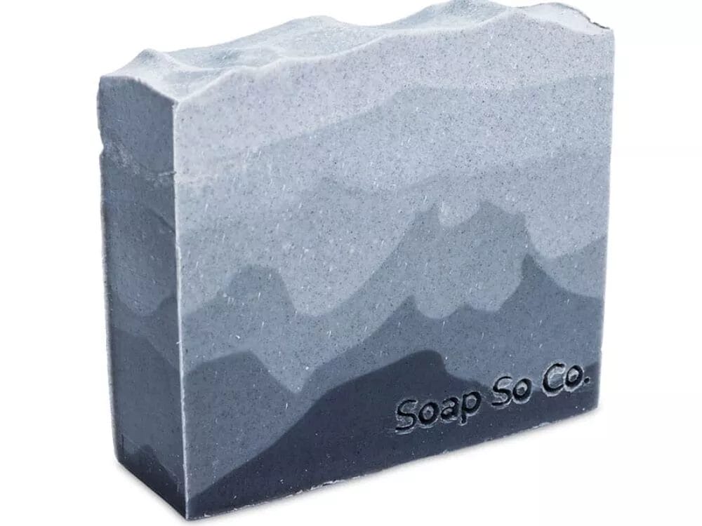 The Soap So Co. bar features a textured pattern reminiscent of mountains, with its surface displaying multiple shades of gray for a layered effect. The brand name is embossed on the lower right corner of the soap.
