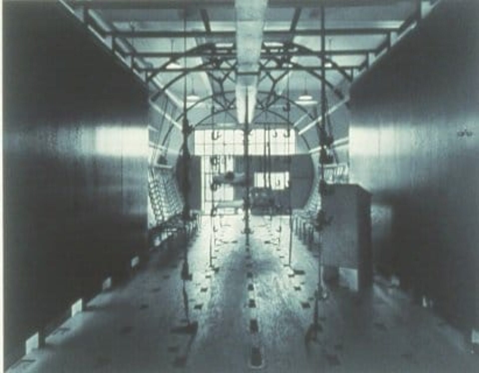 A vintage black and white photo of the interior of an aircraft wind tunnel. The tunnel features large, curved walls and various measuring instruments along its length. Light filters through from the far end, illuminating the space.