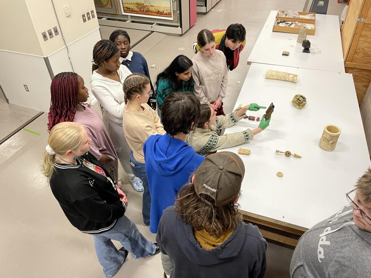 Un groupe de personnes se rassemble autour d'une table dans un atelier ou un cadre éducatif. Une personne fait une démonstration avec un artefact tandis que d'autres regardent attentivement. Divers objets, notamment des pièces sculptées et de la poterie, sont exposés sur la table.
