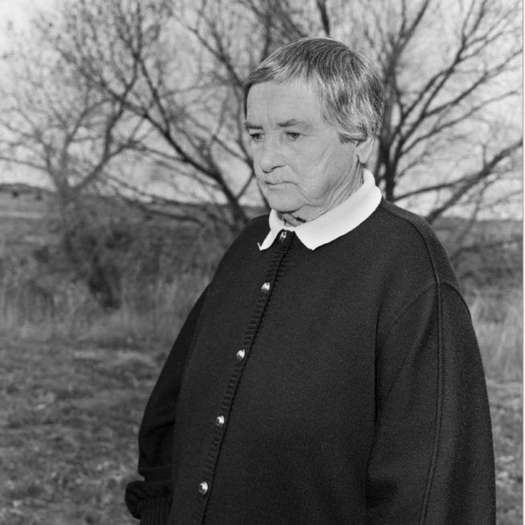 Older man standing facing off to the left with an expressionless face.
