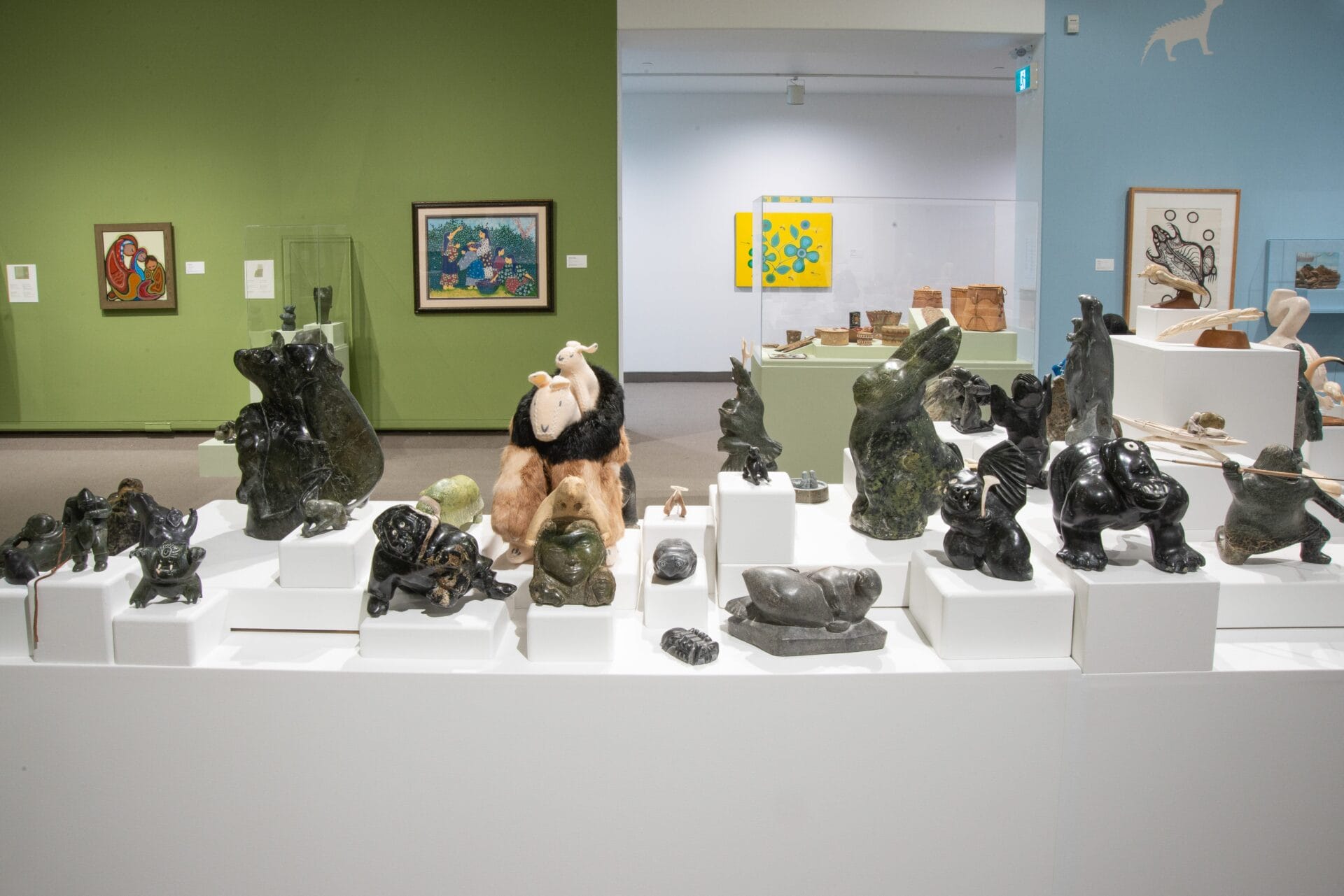 Several Inuit sculptures and carvings are displayed on white pedestals in an art gallery, highlighting the transformation from 2D to 3D. The background features green and white walls with framed paintings and additional art pieces.