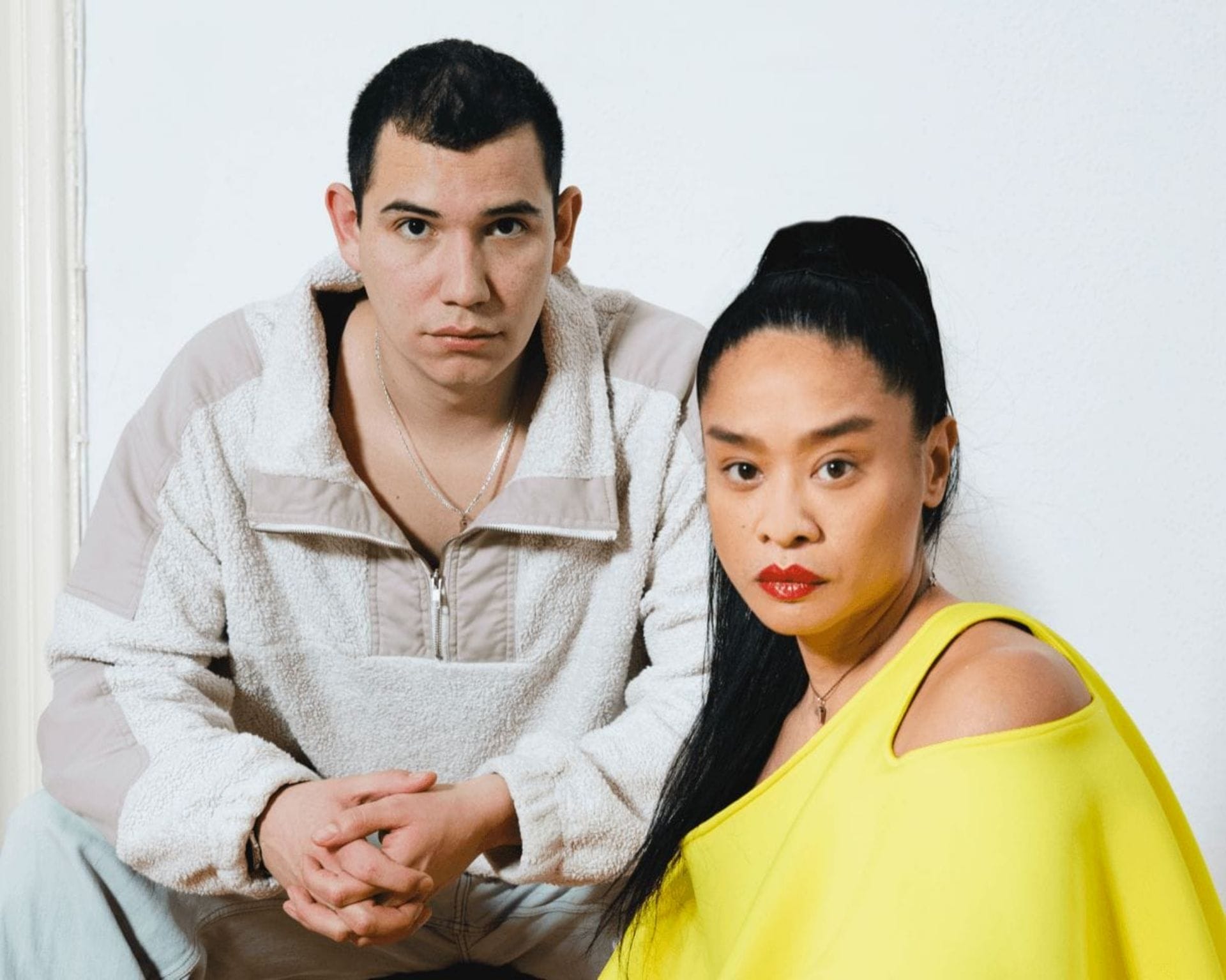 A man and a woman pose against a white background. The man, with short dark hair, wears a light beige fleece pullover and looks directly at the camera. The woman, with long dark hair tied back, wears a bright yellow top with an exposed shoulder, also staring at the camera.