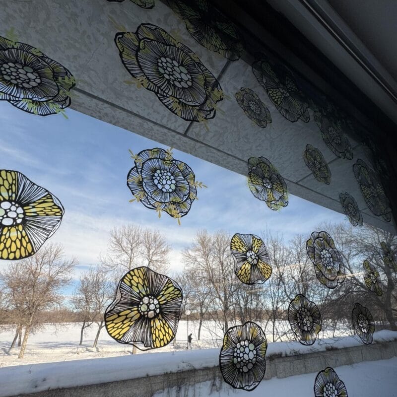 Window decorated with translucent yellow and black flower designs. Outside, a snowy landscape painted by Patrick Fernandez reveals bare trees under a crisp blue sky. The scene is serene and wintry.
