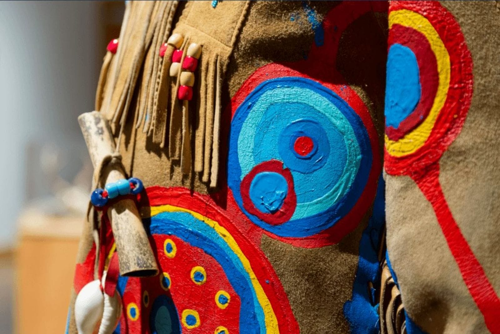 Close-up of a piece of clothing with intricate, colorful designs featuring concentric circles in red, blue, and yellow. The garment includes fringe details and is adorned with beads and a small bone, adding to its elaborate decoration.