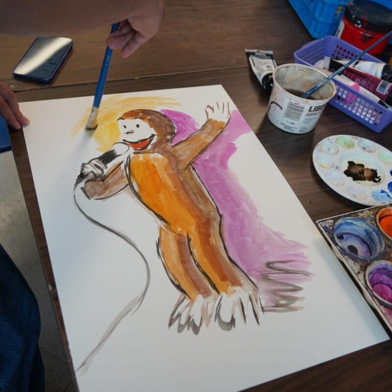 Une personne peint à l'aquarelle sur du papier blanc le portrait coloré d'un singe souriant tenant un micro. Du matériel de peinture et un smartphone sont posés sur la table en bois à proximité.