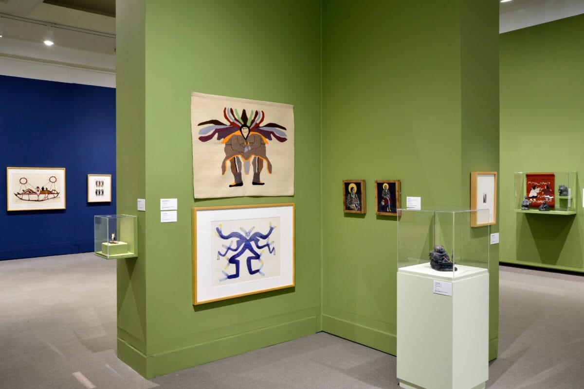 Une galerie de musée aux murs verts et bleus présente des œuvres d'art indigènes colorées, notamment des textiles ornés de motifs d'animaux, des peintures encadrées et de petites sculptures dans des vitrines.