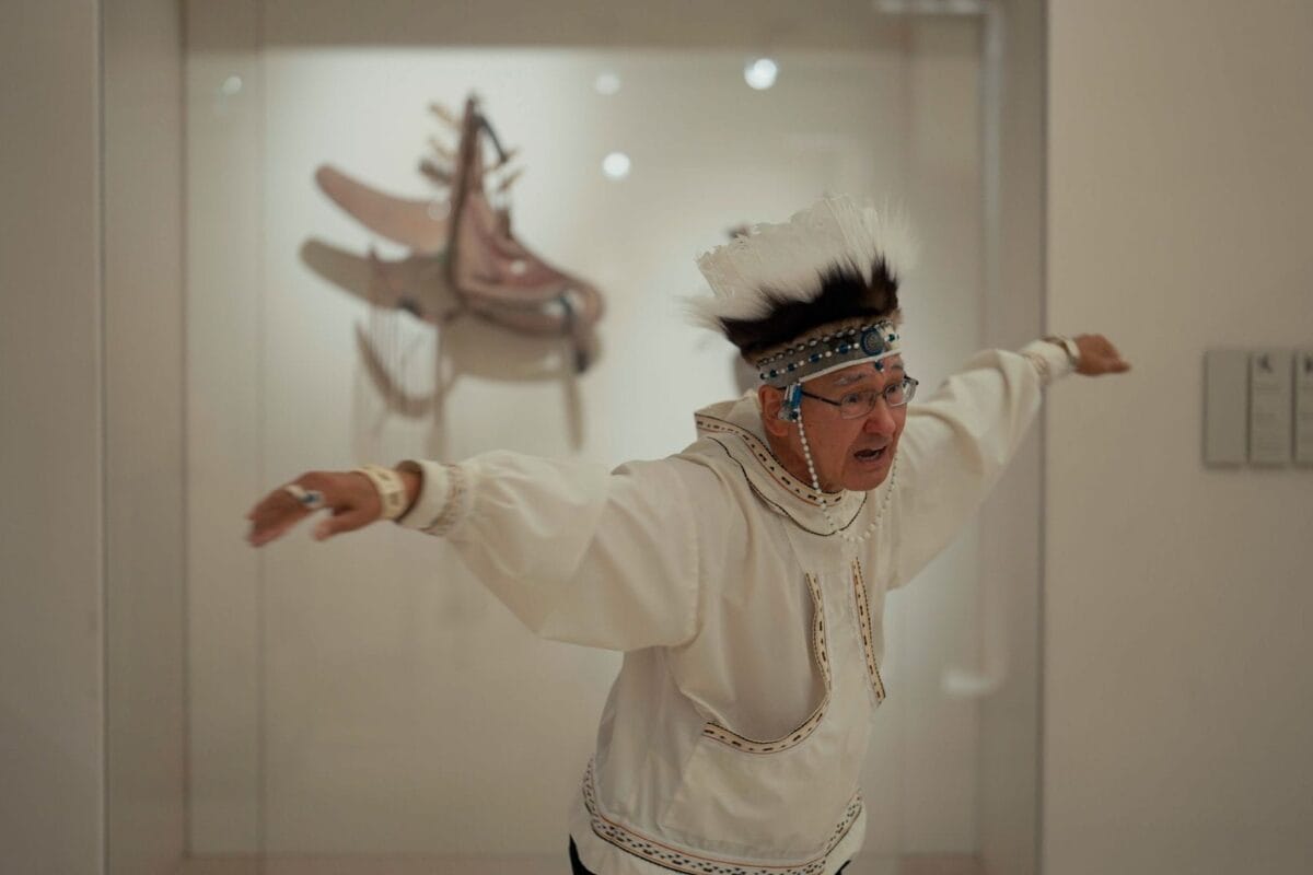 An elder in traditional regalia with a feathered headdress and beaded headband raises his arms outward, appearing to dance, in front of a museum display case containing a saddle—an evocative scene from So Surreal: Behind the Masks.