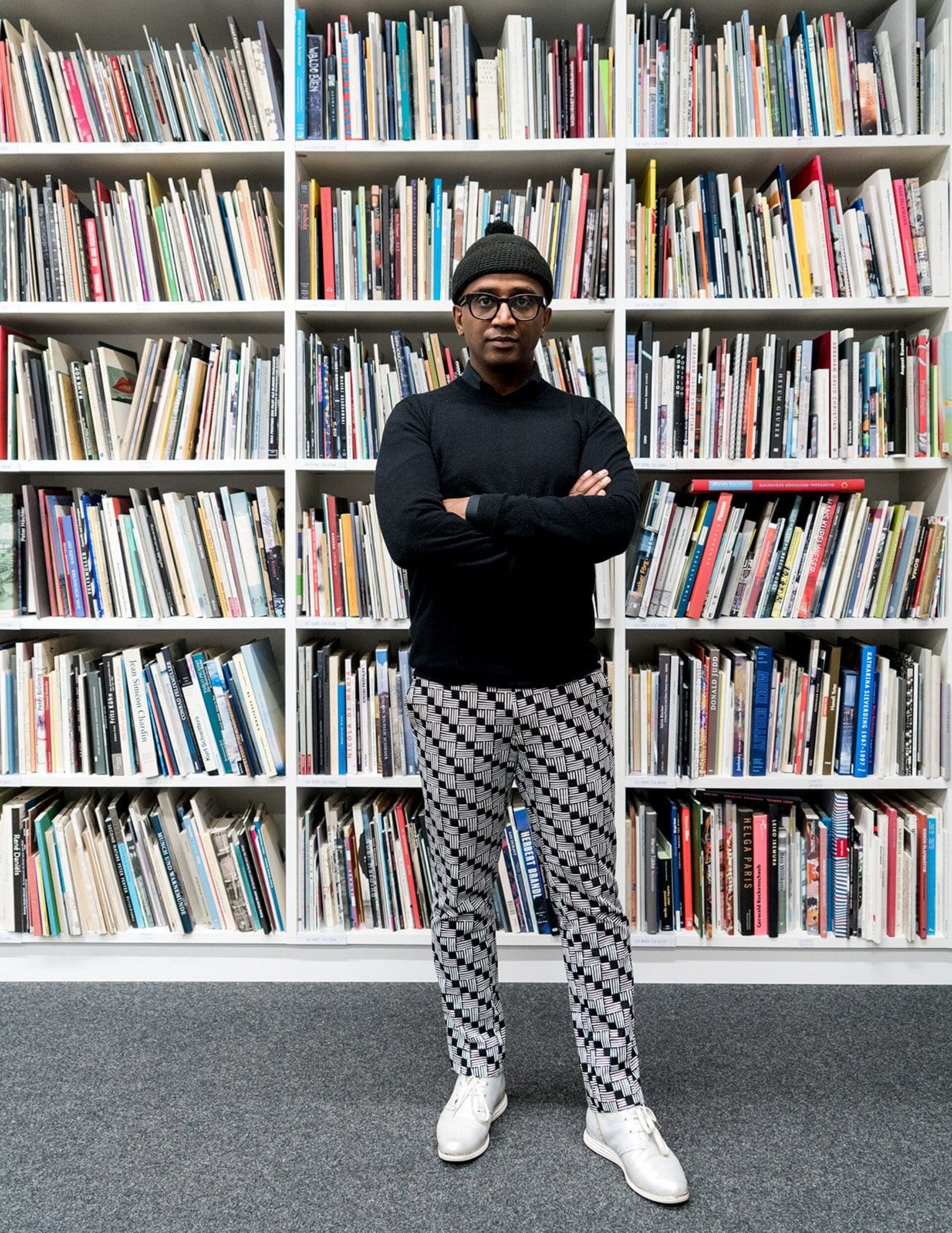 A person wearing a black top, patterned pants, and a black beanie stands with arms crossed in front of a large bookshelf filled with colorful books. The person is also wearing white shoes and glasses.