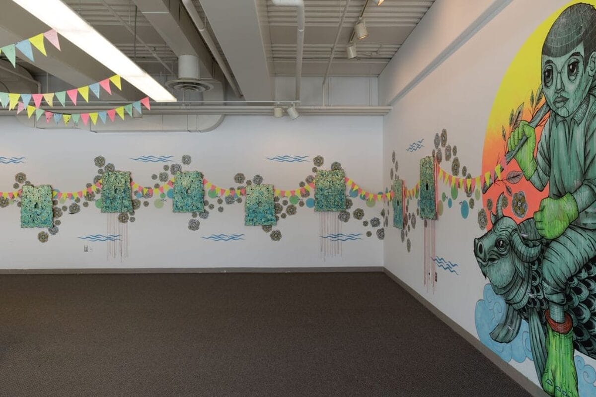 Une pièce spacieuse présente une fresque murale représentant une silhouette chevauchant une tortue sur fond de soleil jaune. Les murs sont ornés d'œuvres d'art abstrait et de guirlandes. Des drapeaux triangulaires colorés sont suspendus au plafond. Le sol est recouvert d'une moquette sombre.