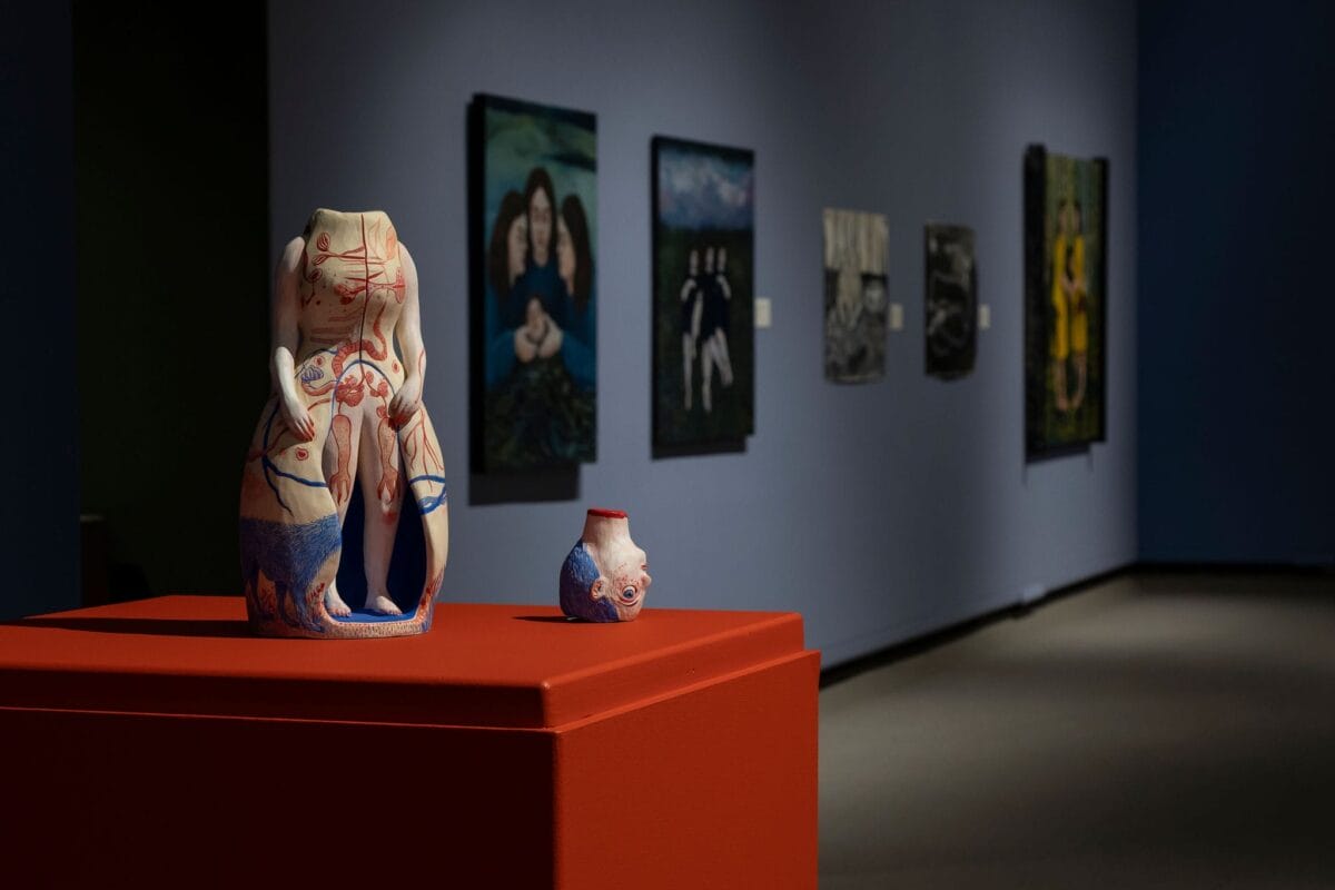 Two abstract, headless ceramic sculptures with anatomical designs are displayed on a red pedestal in a dimly lit art gallery; paintings hang on the dark blue wall in the background.