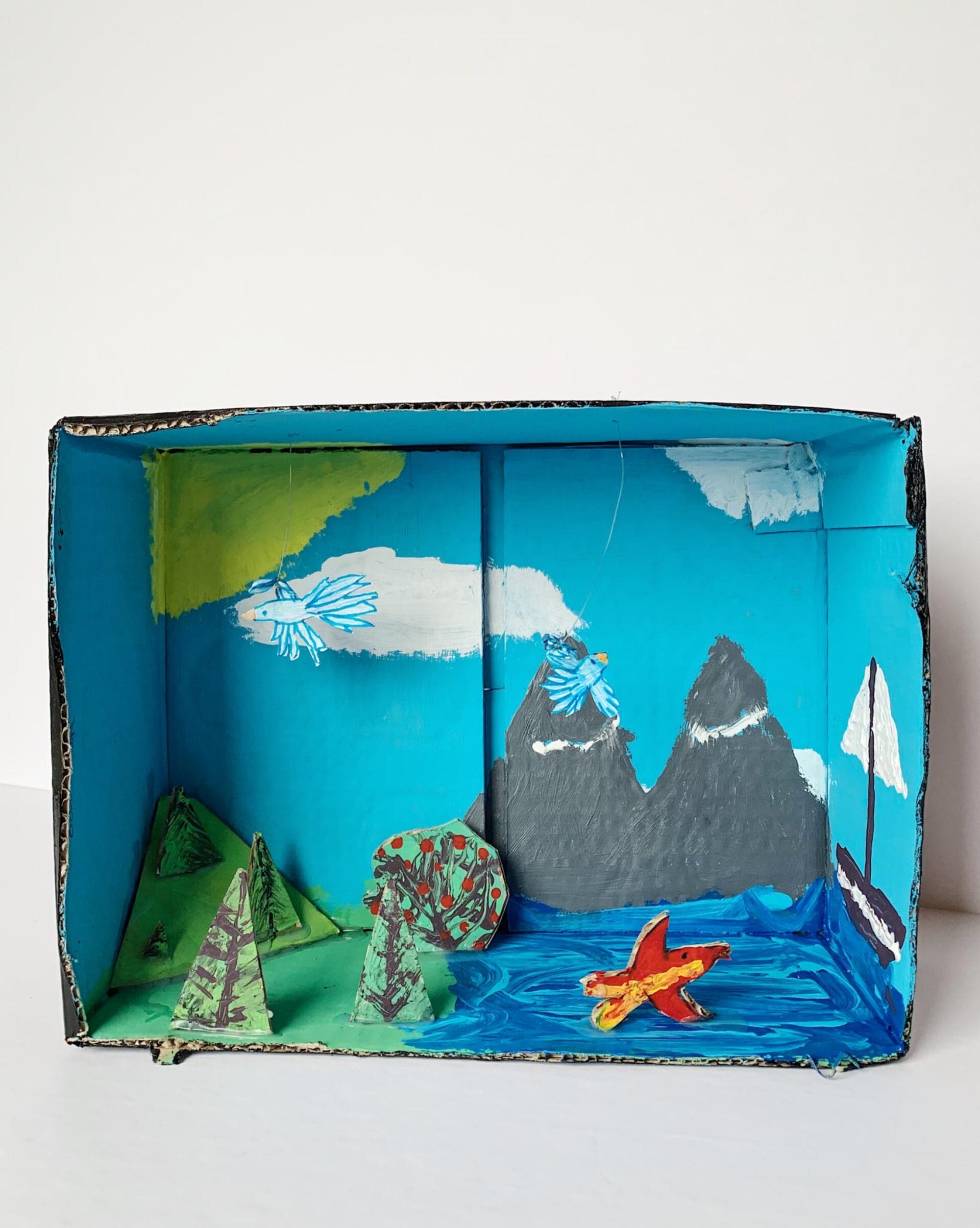 A handmade diorama features a scenic landscape. Mountains, painted mostly black with white peaks, stand against a bright blue sky with fluffy white clouds. The ocean, depicted by a wavy blue base, harbors an orange fish. Trees and hills complete the foreground.