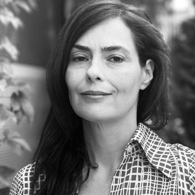 Une femme aux longs cheveux bruns se tient à l'extérieur, vêtue d'une chemise à boutons à motifs. Elle regarde l'appareil photo avec une expression neutre. L'arrière-plan est flou et la verdure est visible. L'image est en noir et blanc.