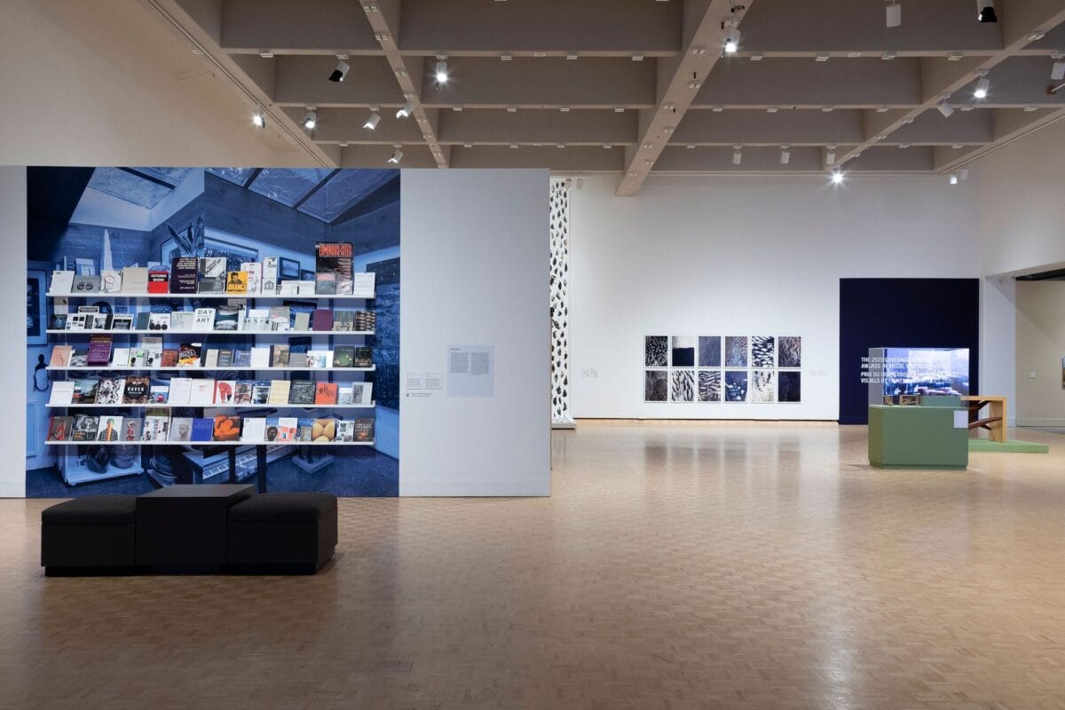 A spacious modern art gallery with parquet flooring, featuring large photo displays on the walls, a bookshelf installation with books, and black benches—spotlights highlight exhibits that celebrate The 2025 Governor General’s Awards in Visual and Media Arts.