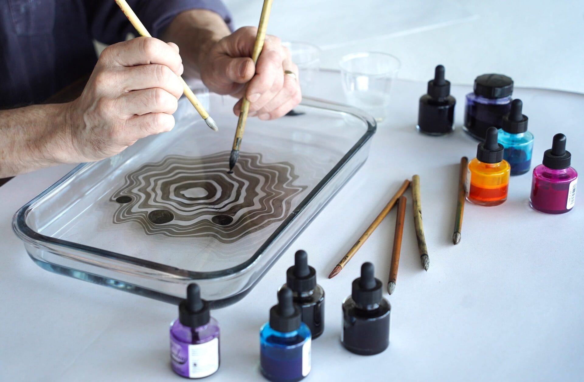 A person uses brushes to create concentric patterns in a glass tray filled with water during classes & workshops, surrounded by bottles of colorful inks and additional brushes on a white surface.