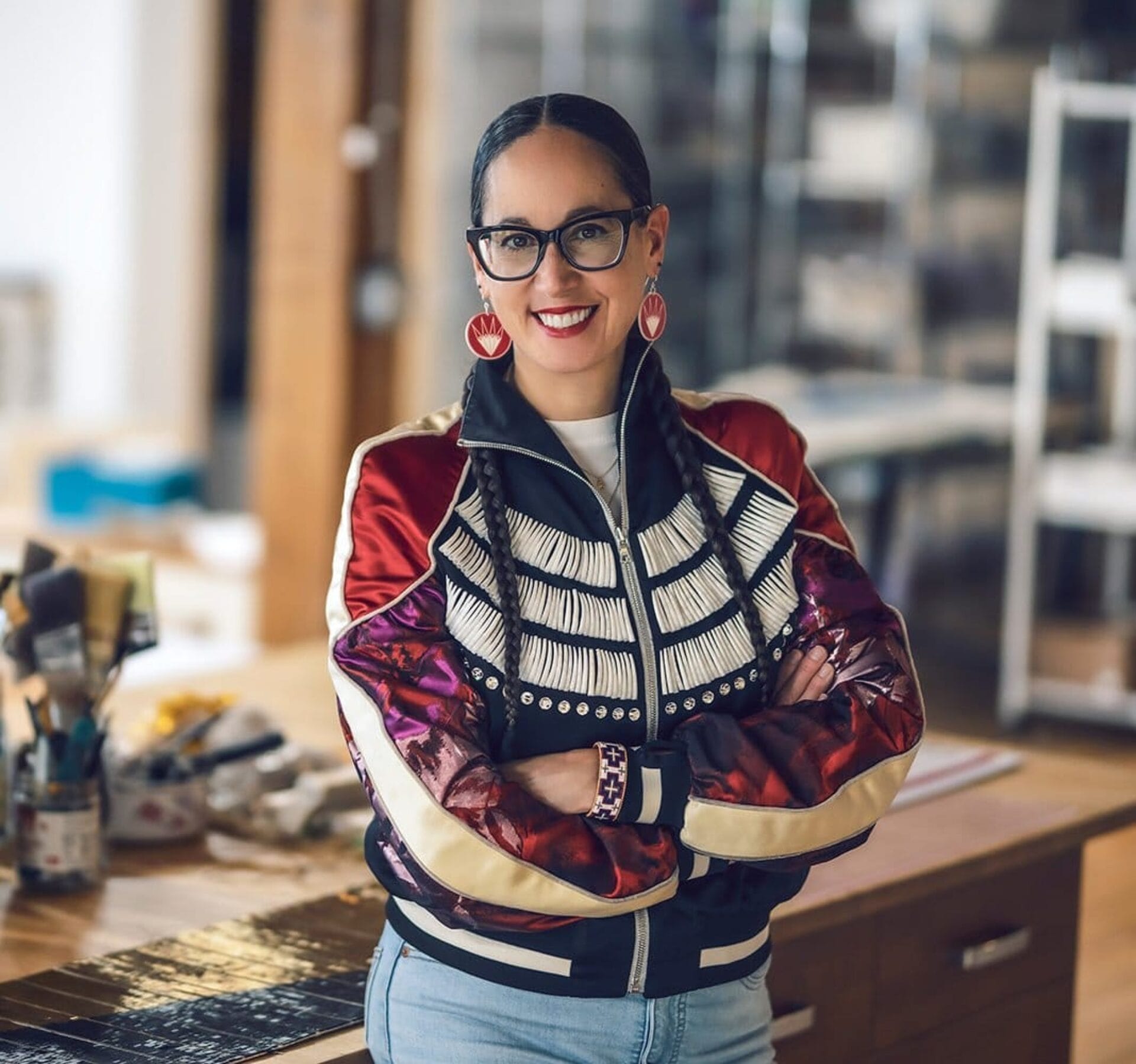 A person with long hair tied back, wearing glasses, a colorful jacket with intricate designs, and statement earrings, standing with arms folded in a craft room. The background features art supplies and a wooden table. The person is smiling.