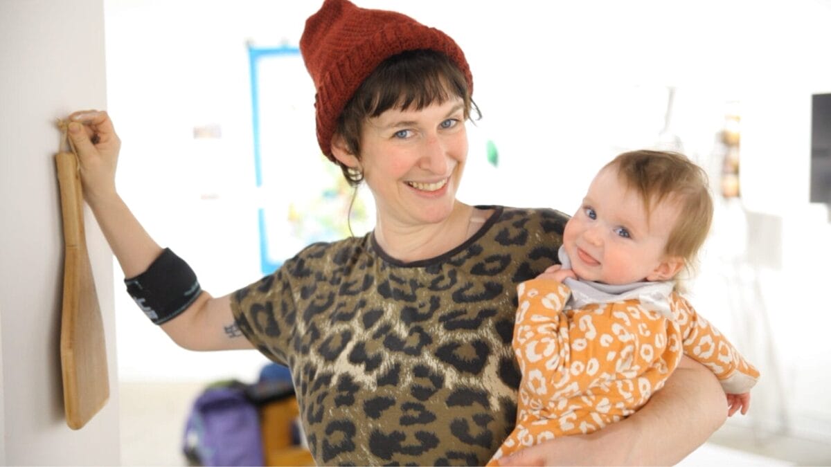 Une femme souriante, coiffée d'un bonnet en tricot rouge et vêtue d'une chemise imprimée léopard, tient dans ses bras un bébé heureux vêtu d'une grenouillère à motifs orange, à l'intérieur d'une pièce lumineuse. Le bras gauche de la femme repose sur un objet en bois fixé au mur.