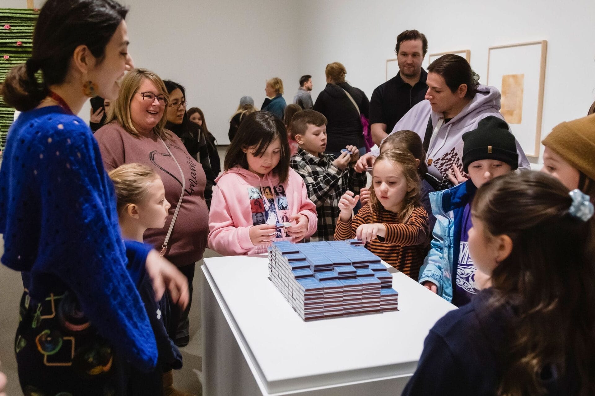 A group of children and adults gather around a display table with stacked blue blocks in an art gallery, engaging with the exhibit while observing and interacting with the objects.