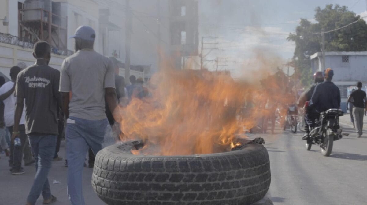 Protest scene with a large tire burning on a city street, emitting smoke. People walk by, and a motorcyclist rides past. The background shows buildings under a clear sky.