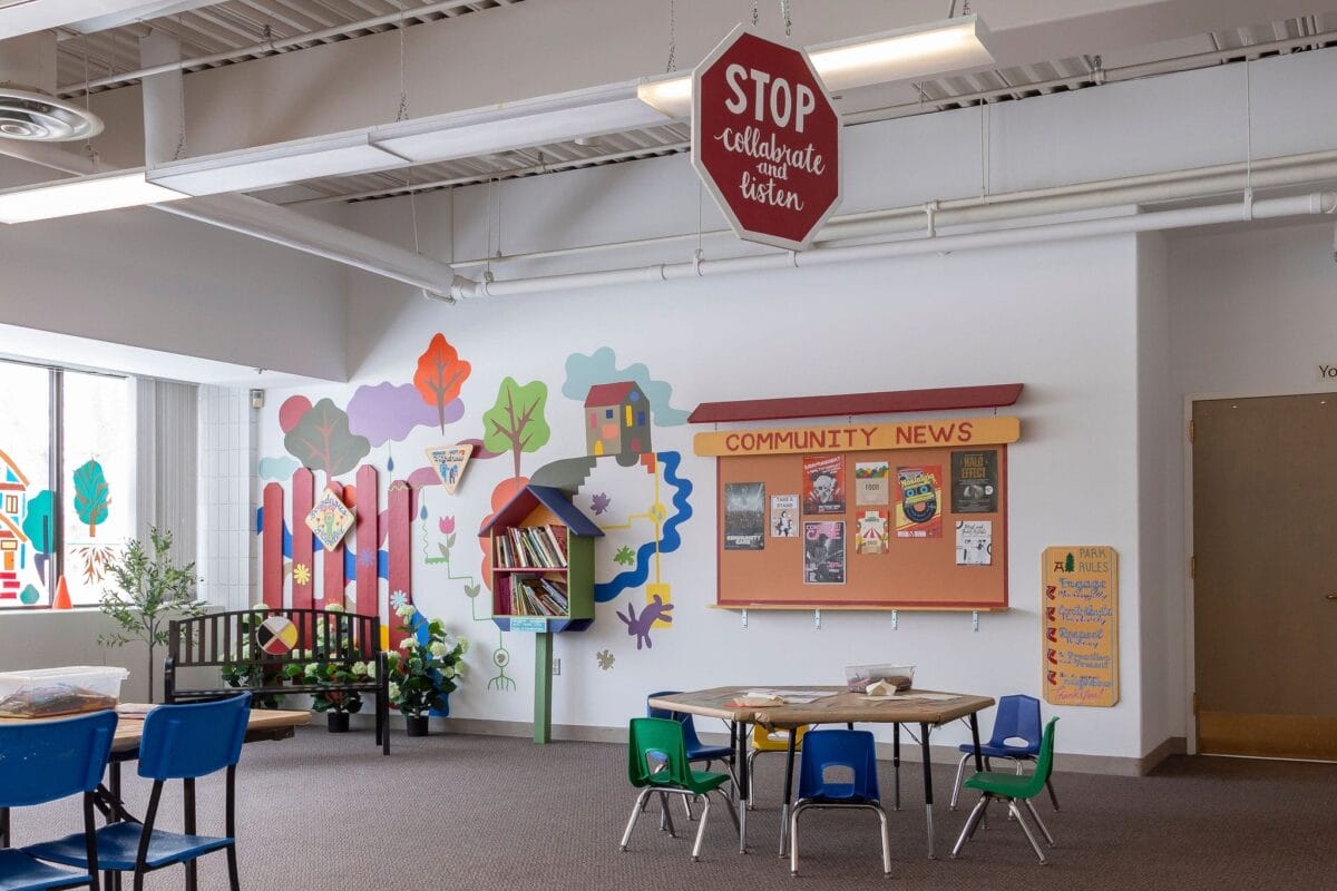 Une salle de classe colorée avec une fresque d'arbres et de maisons, une petite bibliothèque, un tableau d'affichage intitulé 