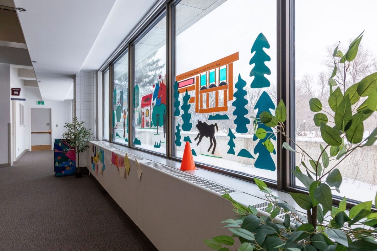 Un couloir d'école avec de grandes fenêtres décorées d'animaux colorés et de découpes de la nature. La neige est visible à l'extérieur. Des plantes vertes sont disposées le long du couloir et un cône orange vif est posé sur le rebord de la fenêtre.