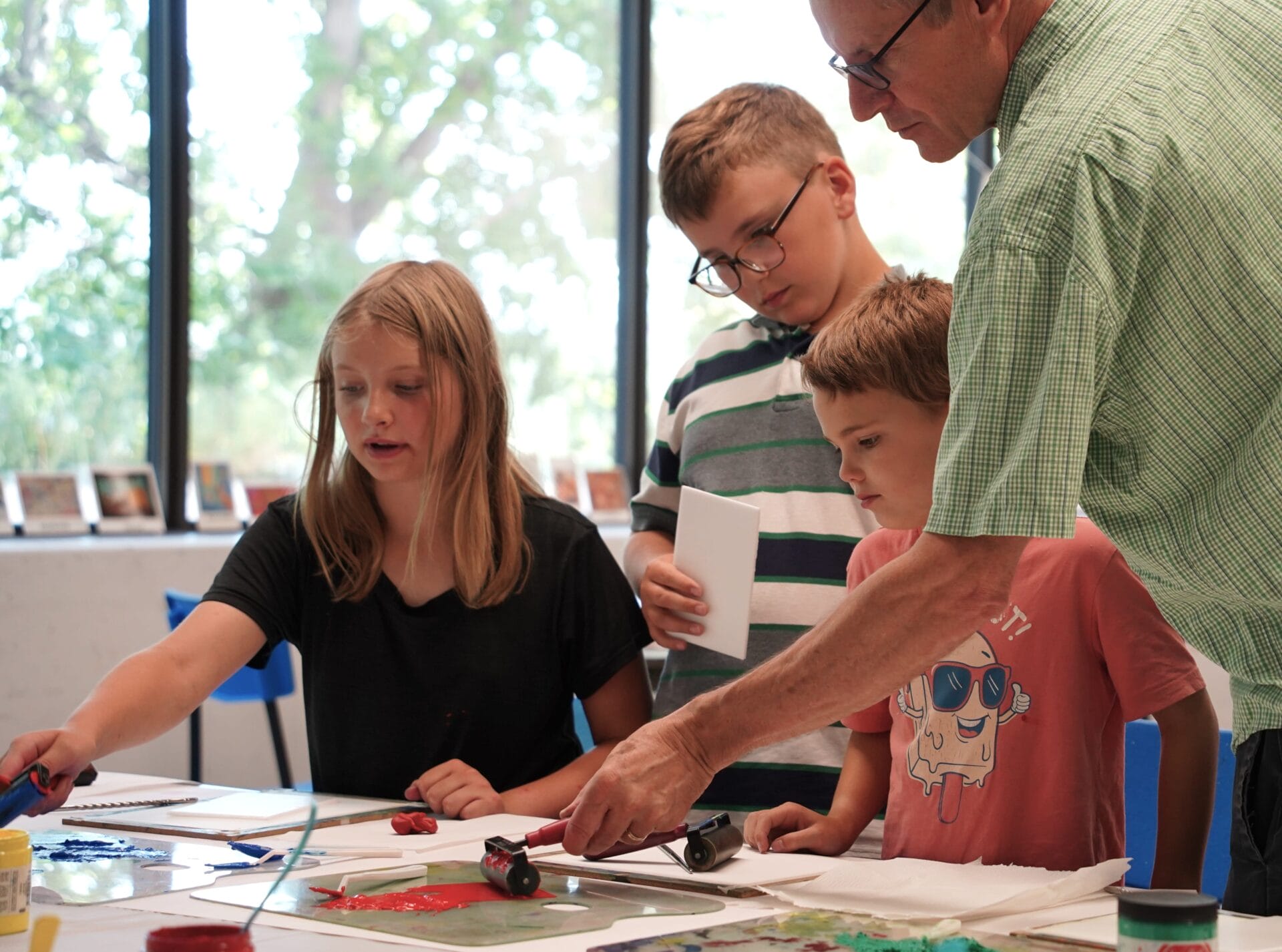 A man leads three children in an art project at a table covered in paper, paint, and tools. The children watch and participate, reflecting the creative spirit of Classes & Workshops, with large windows and greenery visible in the background.