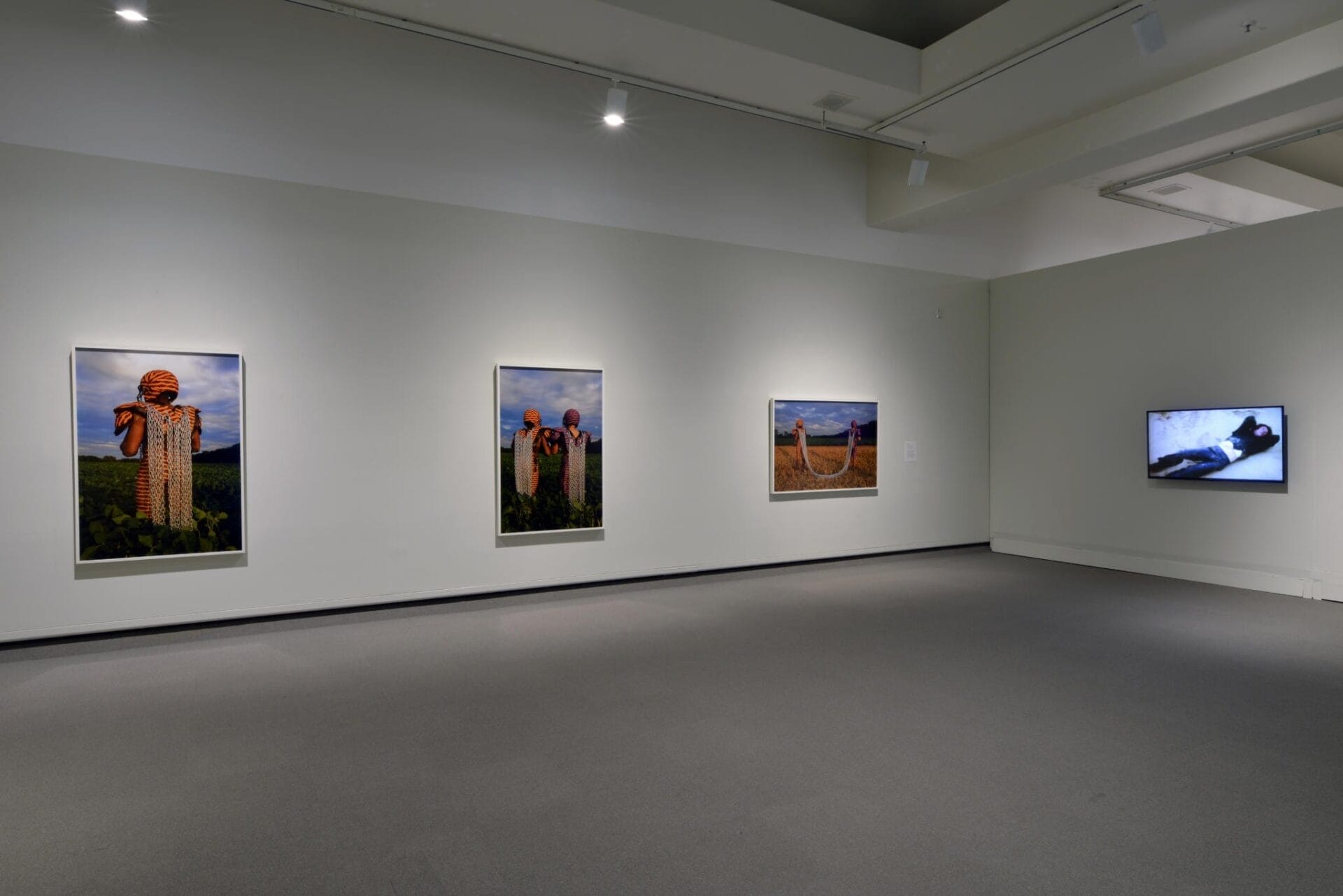 A modern art gallery features three large photographs of figures in elaborate attire on a white wall, with a fourth black-and-white image displayed on a monitor to the right. The room has grey carpeting and soft overhead lighting.