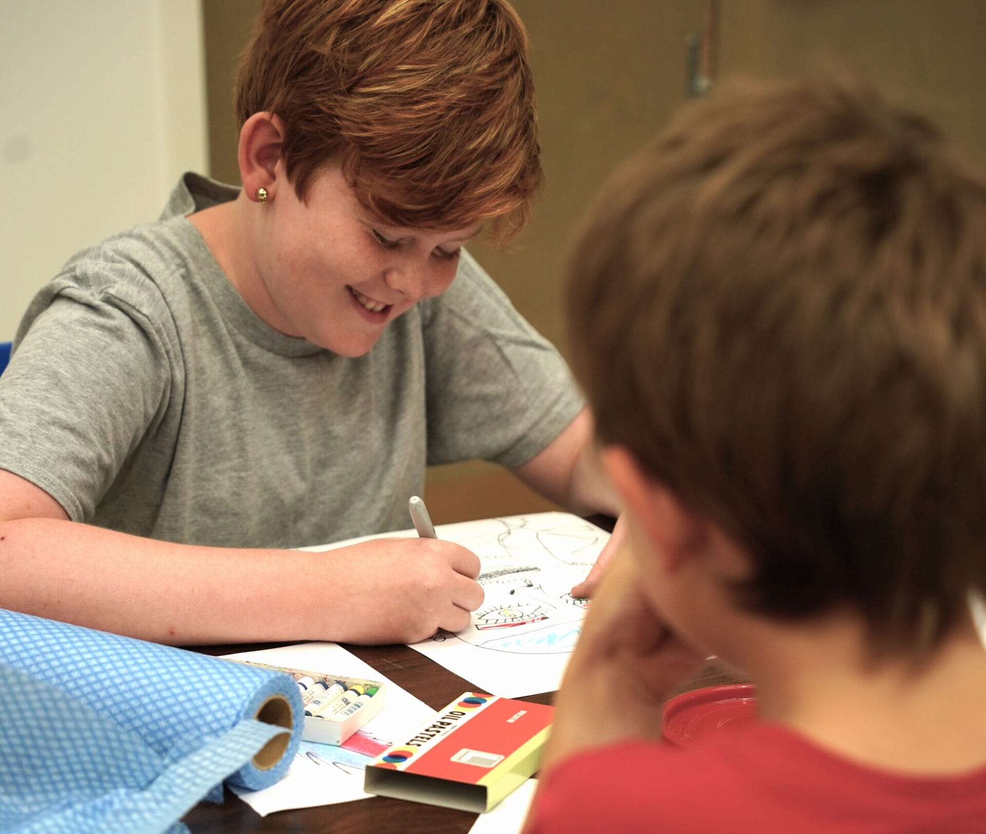 Two children sit at a table drawing with markers during one of our creative classes & workshops. One child with short red hair is smiling while working on their drawing. Various art supplies and papers are spread out on the table.