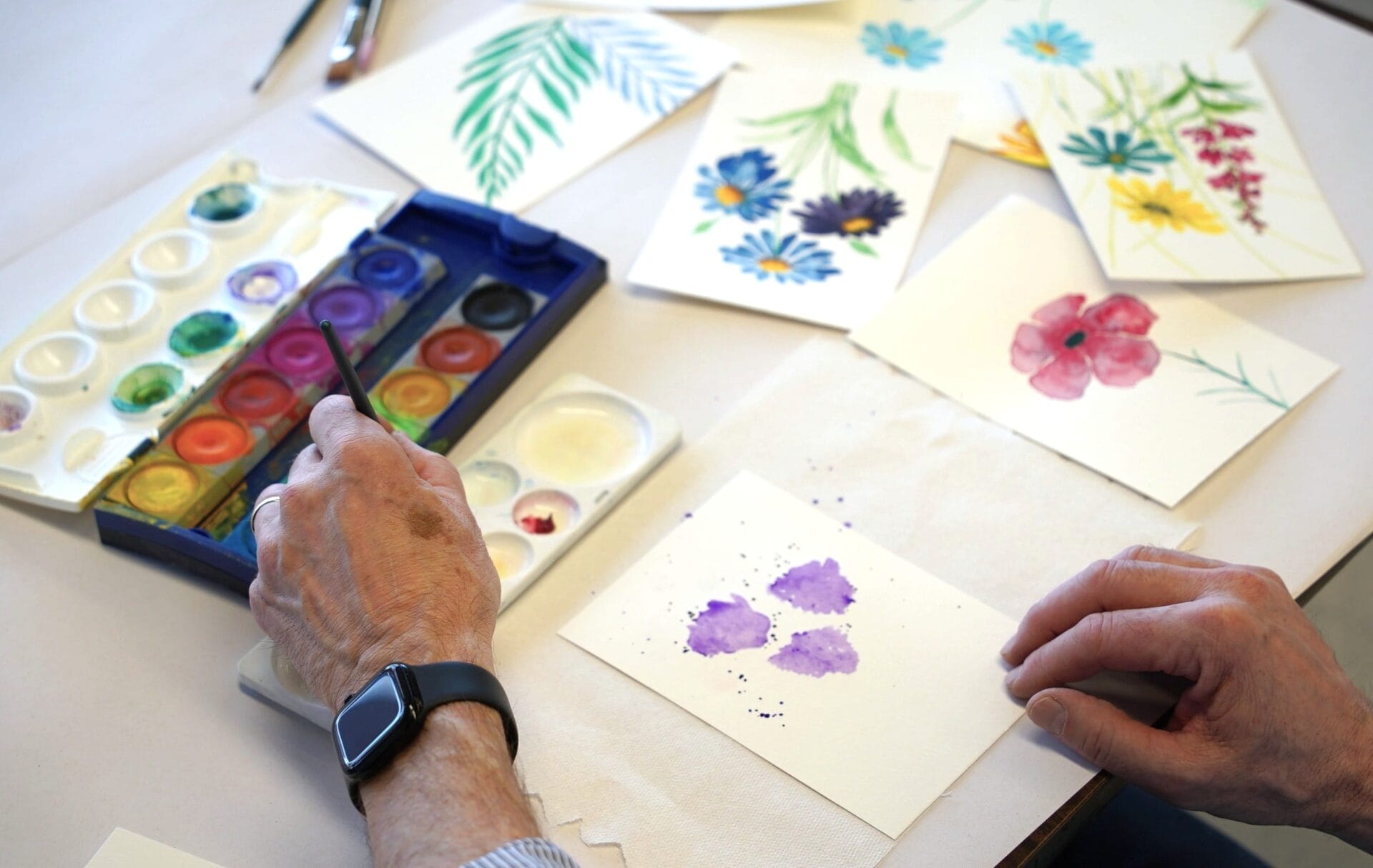 A person with a smartwatch paints purple flowers on paper using watercolors during Classes & Workshops. Several finished floral watercolor paintings and a paint palette are spread out on the table.