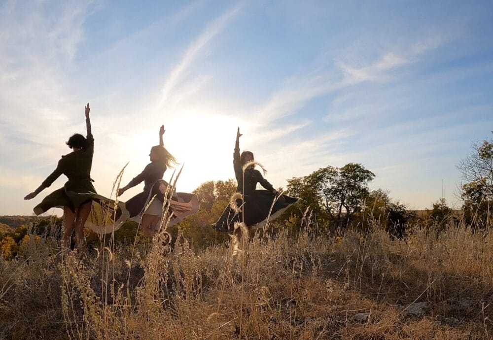 Four people in dresses leap into the air with arms raised in a grassy field at sunset, with tall dry grass and trees in the background, silhouetted against a bright sky.