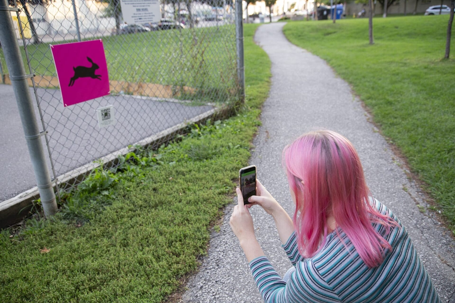 Une personne aux cheveux roses est agenouillée sur un sentier et prend en photo avec son téléphone un panneau rose orné de la silhouette d'un lapin noir sur une clôture grillagée. Le sentier serpente vers une zone herbeuse avec des arbres et des bâtiments en arrière-plan.