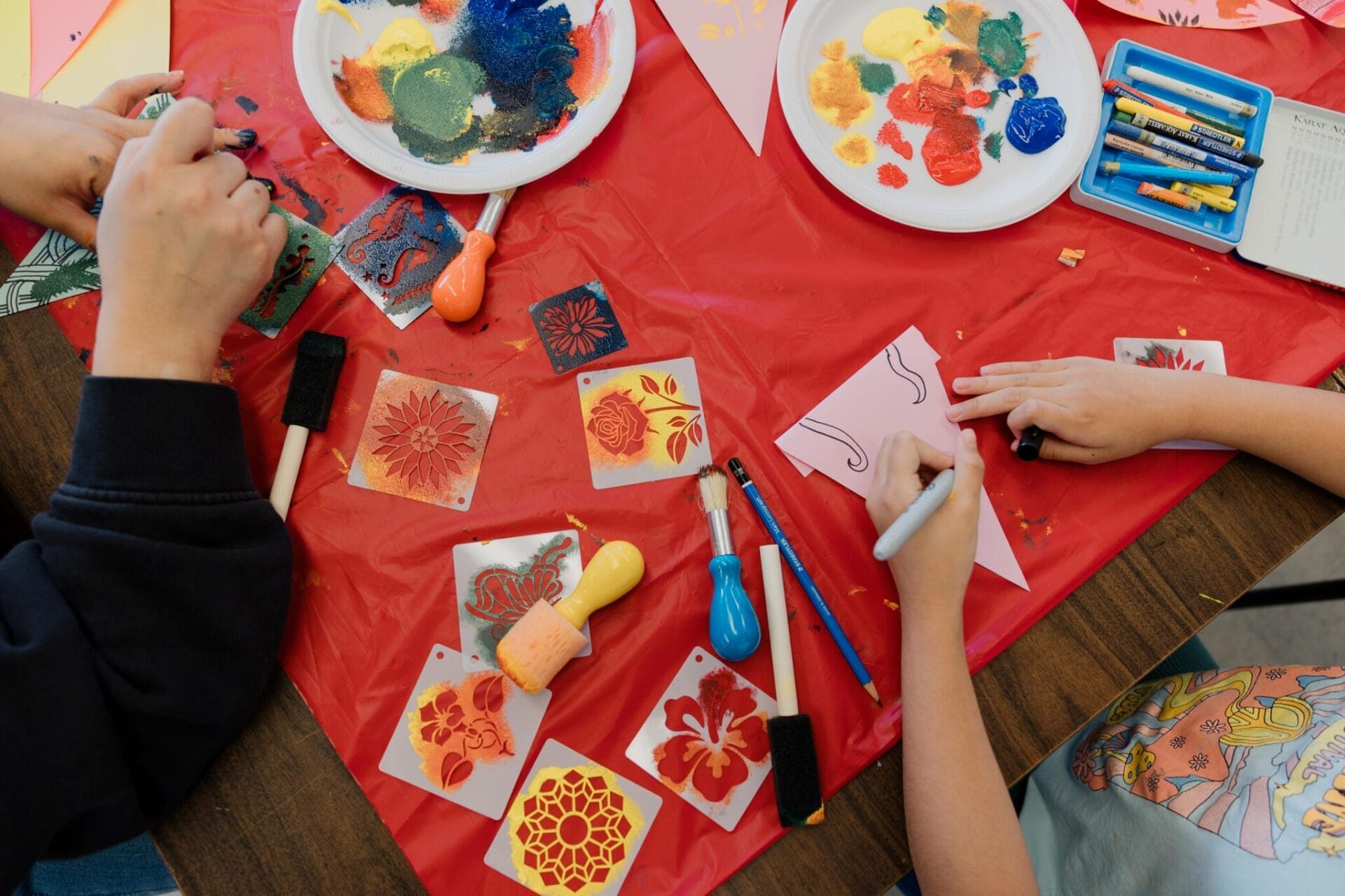 Deux personnes utilisent des marqueurs, de la peinture et des tampons pour créer des œuvres florales sur papier, assises sur une table recouverte d'une nappe en plastique rouge. Divers pochoirs, assiettes recouvertes de peinture et outils de bricolage sont disséminés un peu partout.