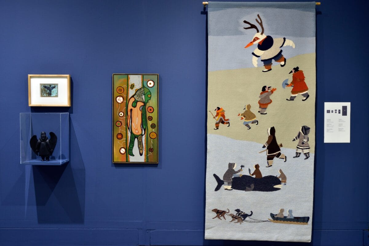 Sur un mur bleu, un musée expose de l'art inuit : une petite gravure encadrée, une sculpture sombre dans un coffret, une peinture colorée et une grande tapisserie représentant des personnages en costume traditionnel avec des animaux et des scènes ludiques.