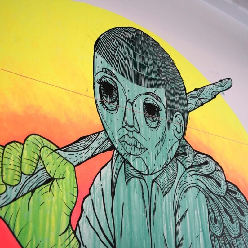 A vibrant mural by Patrick Fernandez depicts a figure with a stylized face and large eyes, holding a stick over their shoulder. The background features a gradient of yellow to orange, creating a vivid contrast with the green and black details of the figure.