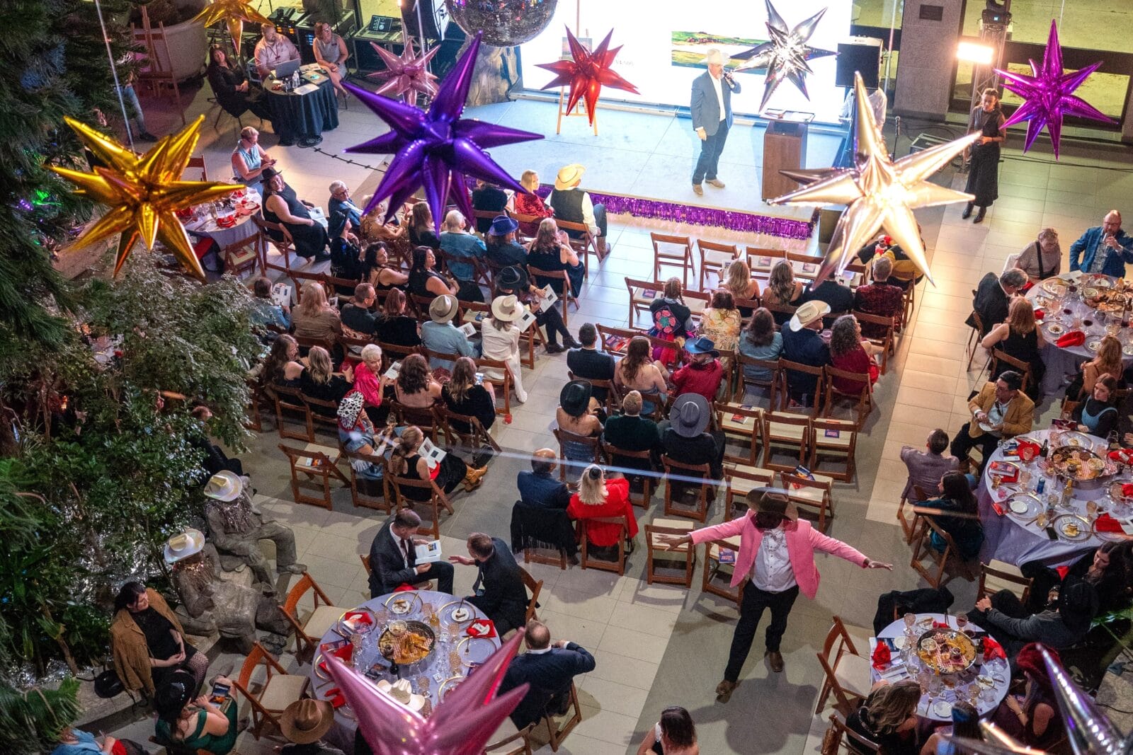 A lively indoor event with people seated at round tables, some wearing hats. Colorful star-shaped decorations hang from the ceiling. A person in a pink jacket stands, while someone speaks on stage under bright lights.