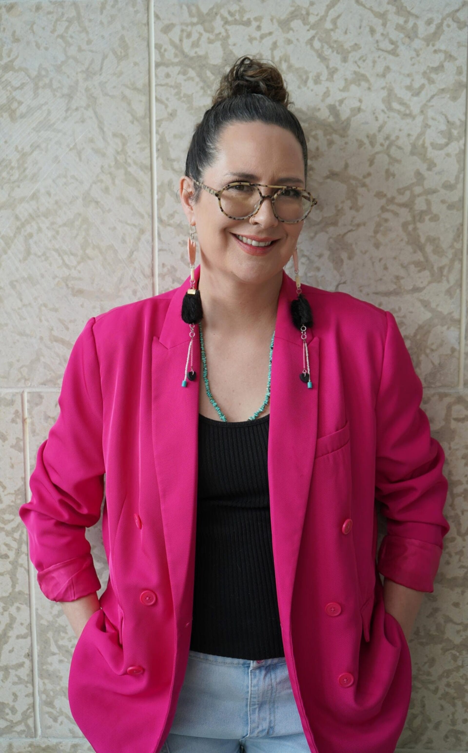 A person with a topknot hairstyle smiles while standing against a beige textured wall. They wear glasses, long beaded earrings, a bright pink blazer, and a black top with light blue jeans. Their hands are casually placed in their pockets.