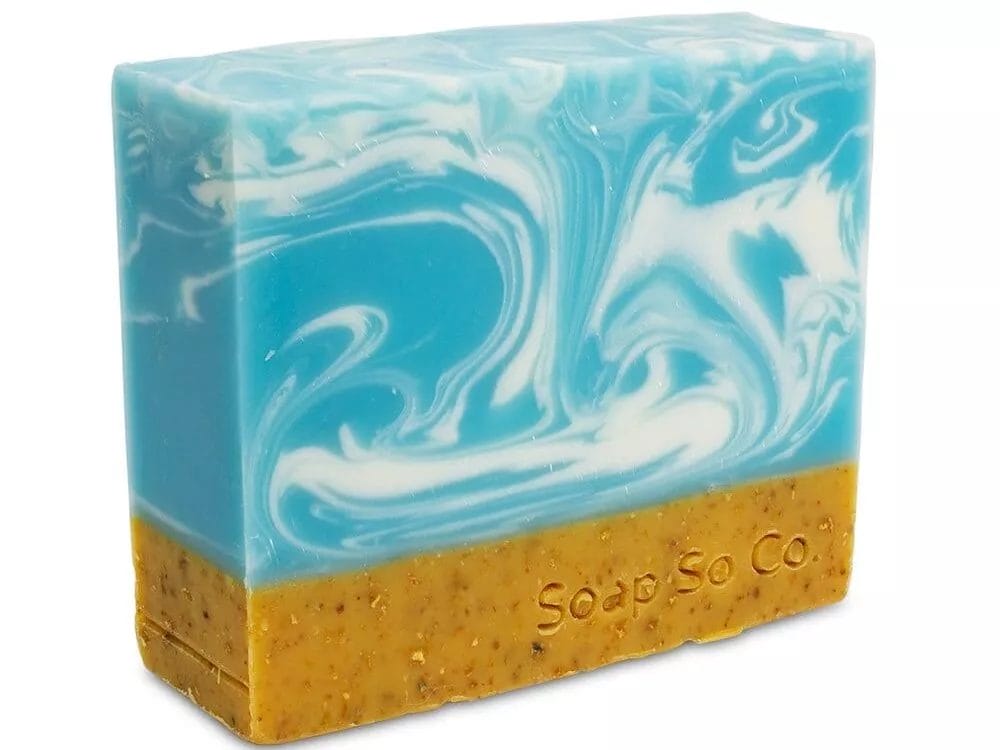 A handcrafted Soap So Co. bar features upper blue and white swirls reminiscent of waves, while the lower brown section has a grainy texture with 